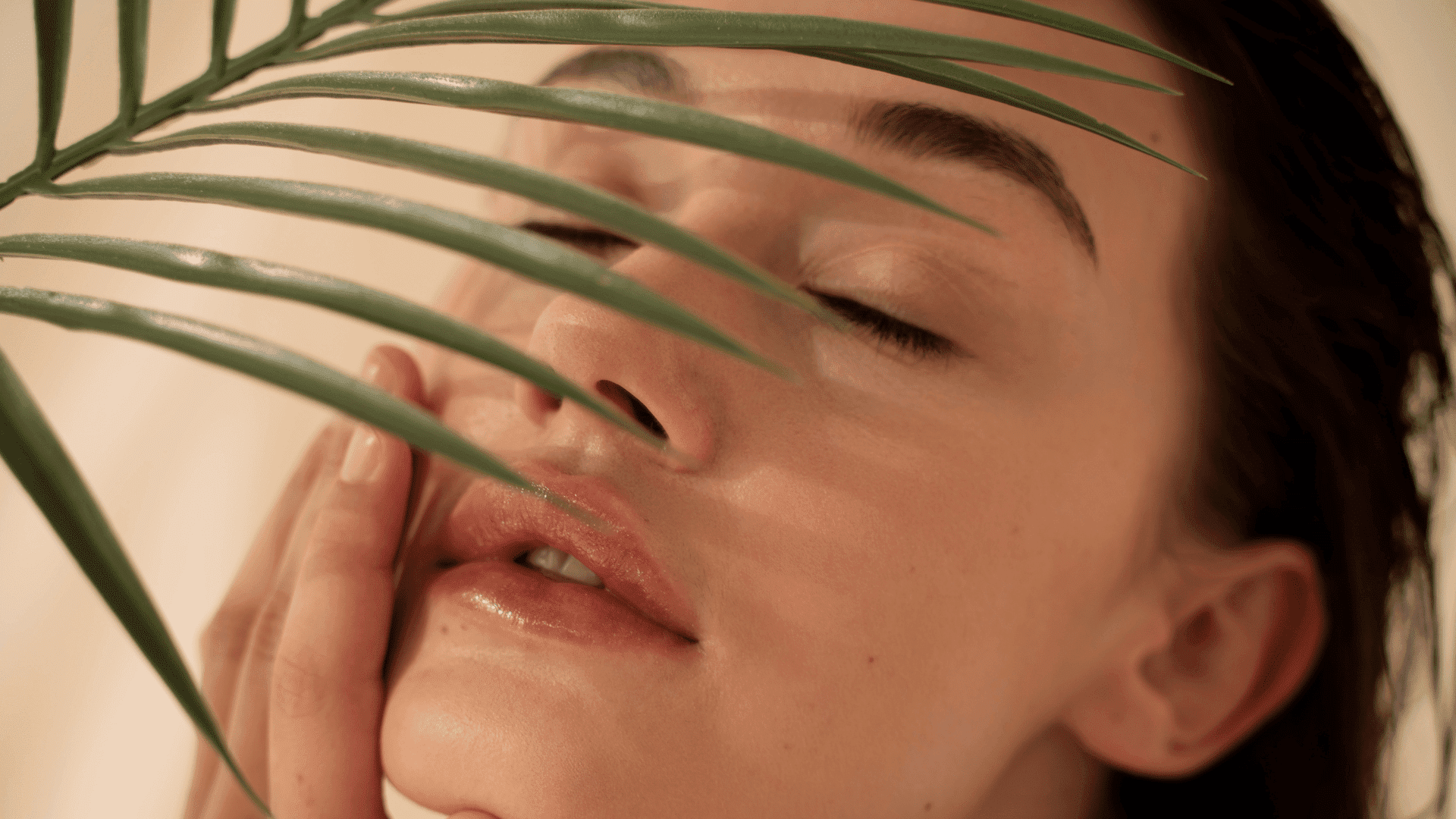 A person with closed eyes gently touching their face, partially covered by a palm leaf.